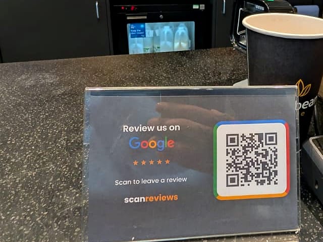 A ScanReviews Google review card on the coffee bar of a forecourt cafe below the drinks menu