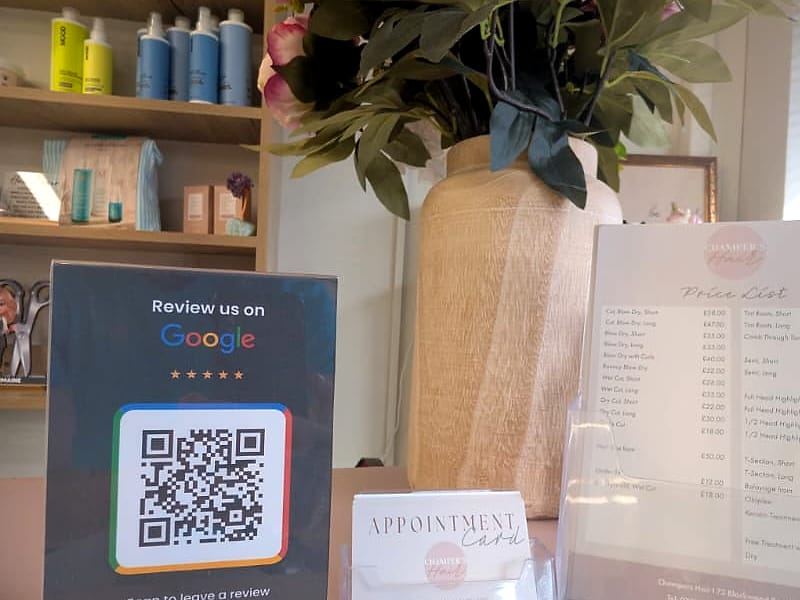 A ScanReviews Google review card in its acrylic stand on a hair salon front desk, beside the appointment cards and price list